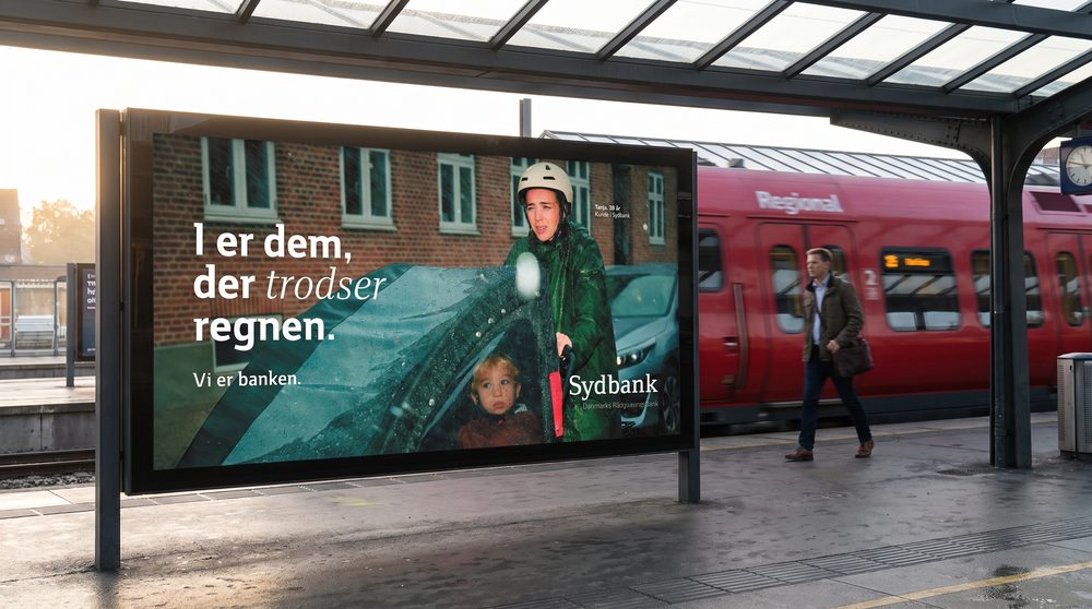 Sydbank-railboard på dansk perron i morgenlys, rødt tog passerer