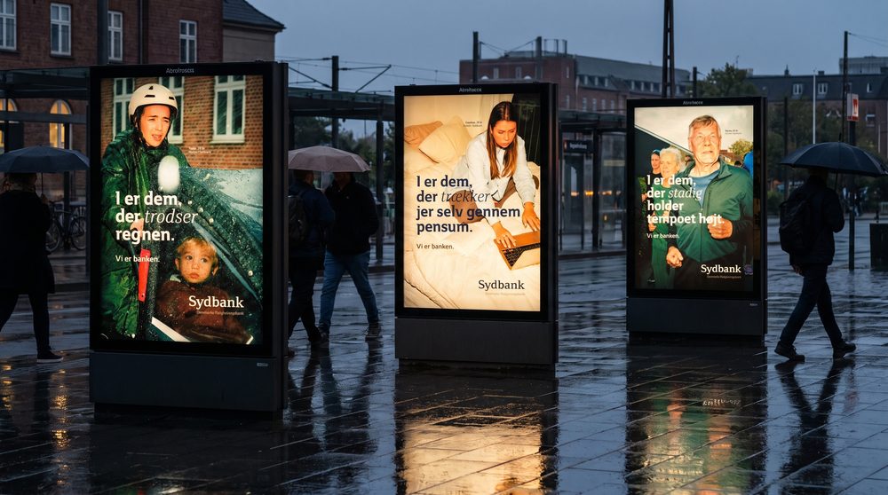Tre Sydbank-lyskasser i række på en regnvåd plads i skumringen